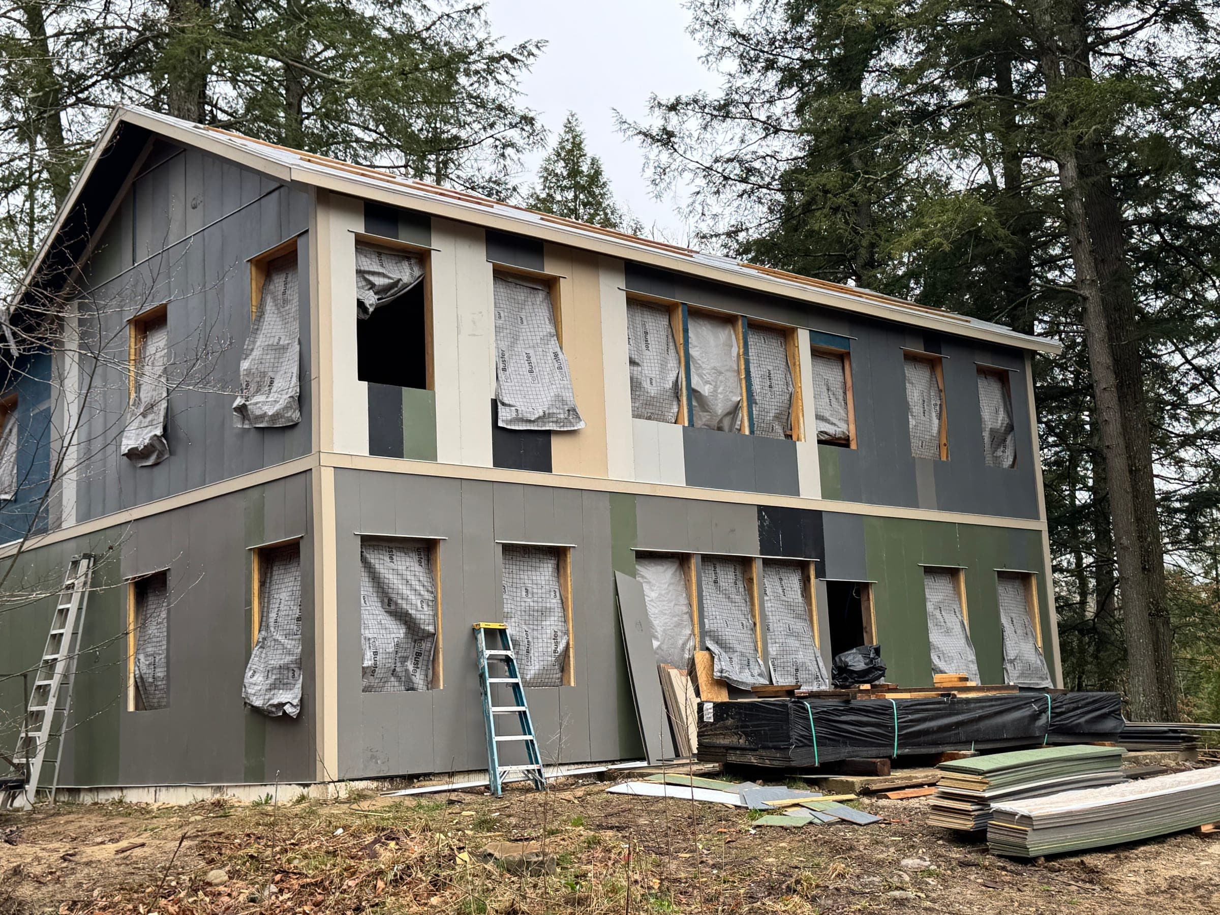 Surplus siding going up on a two-story wall with the factory colors still mixed — gray, cream, olive and charcoal boards side by side
